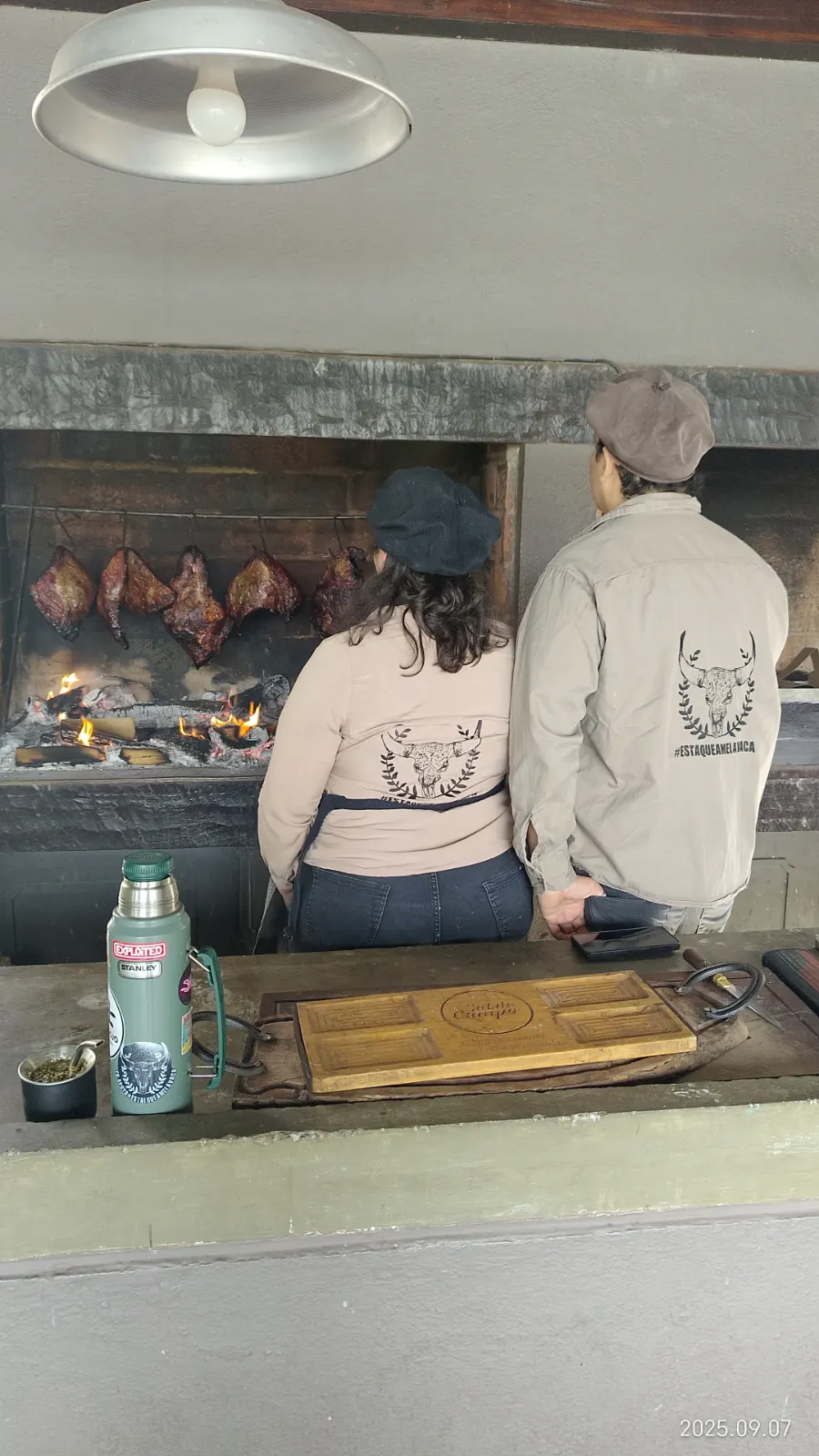 Agustina y Josué de espaldas, con camisas de la marca, frente al fuego